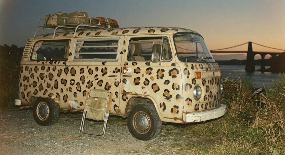 A VW camper van decorated in leopard spots with Menai Bridge in the background
