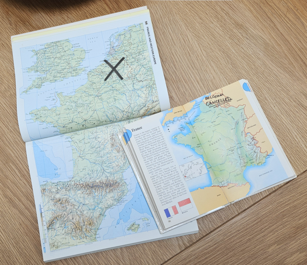 Two atlases on a table open at pages with maps of Europe. On one, there is a cross over country of Belgium. On the other it says, "Belgium Cancelled".