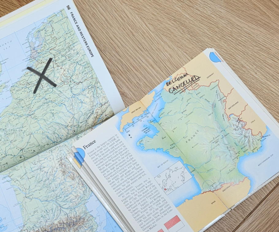 Two atlases on a table open at pages with maps of Europe. On one, there is a cross over country of Belgium. On the other it says, "Belgium Cancelled".
